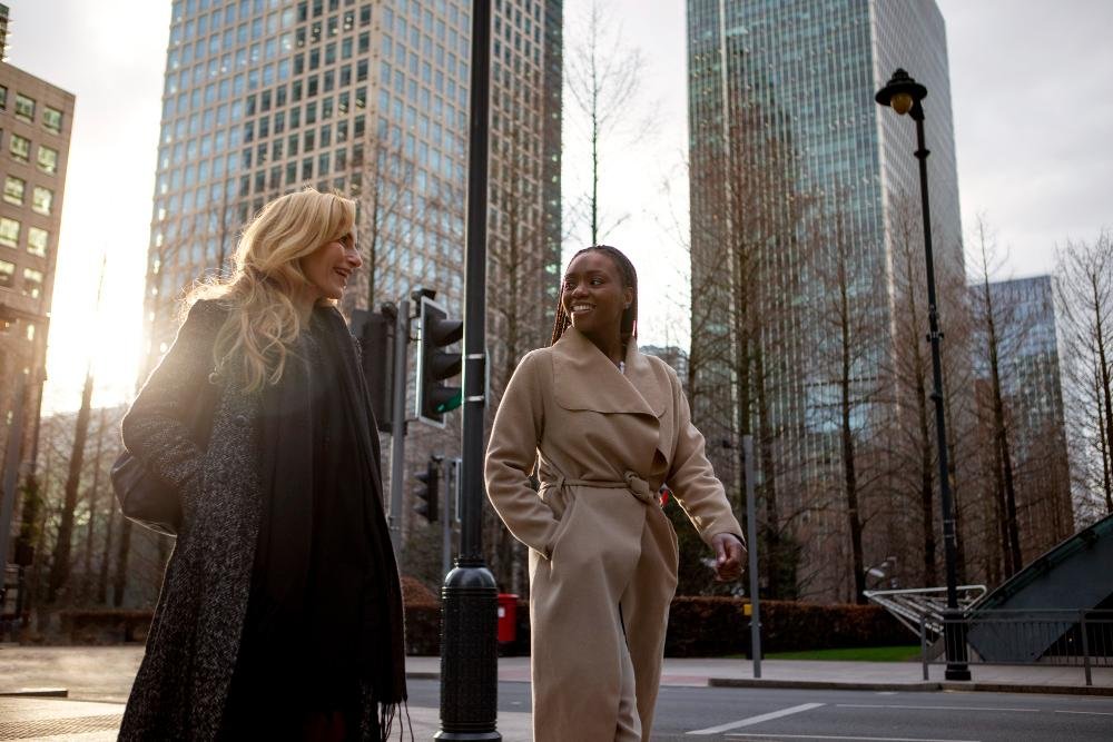Duas mulheres caminhando e conversando em uma rua urbana, com pr&eacute;dios comerciais altos ao fundo, em um ambiente corporativo moderno