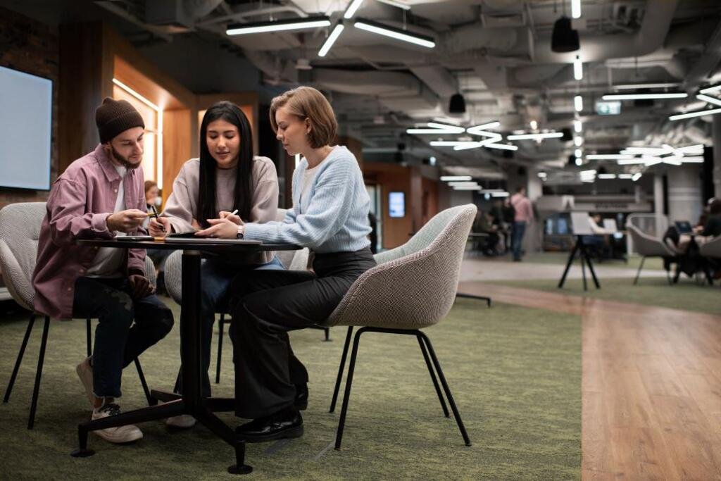Imagem de tr&ecirc;s pessoas jovens em um ambiente moderno de coworking, discutindo e olhando para o celular, com ilumina&ccedil;&atilde;o sofisticada e atmosfera colaborativa.
