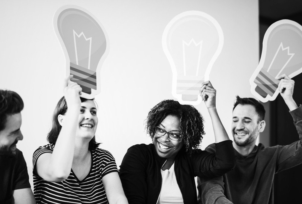 Grupo de pessoas felizes segurando l&acirc;mpadas de desenho, simbolizando ideias criativas e inova&ccedil;&atilde;o em um ambiente colaborativo.