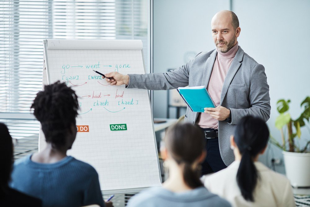 Homem apresentando informa&ccedil;&otilde;es de ingl&ecirc;s para neg&oacute;cios enquanto aponta para anota&ccedil;&otilde;es sobre verbos no flip chart.