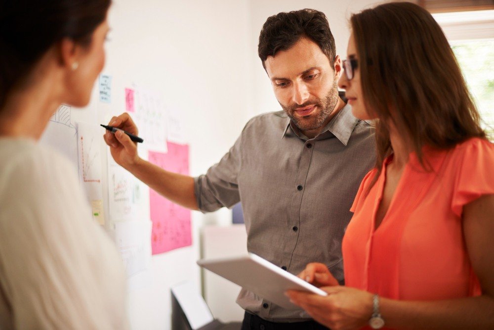 Equipe em reuni&atilde;o analisando gr&aacute;ficos e post-its em um quadro, focada no desenvolvimento de compet&ecirc;ncias no ambiente corporativo.